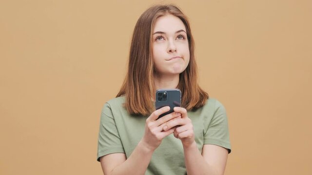 A thinking young woman is using her smartphone standing isolated over a beige wall in the studio