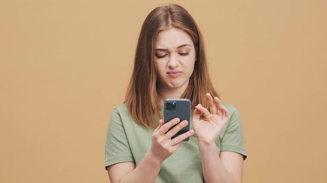 A pretty nice young woman is using her smartphone standing isolated over a beige wall in the studio
