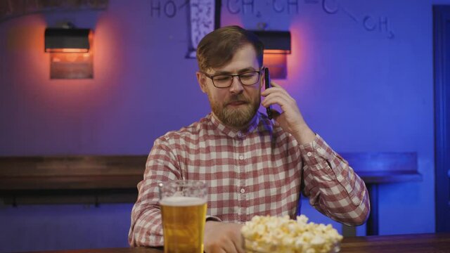 Portrait of handsome man with beard and in glasses making mobile phone call in pub. happy millenial ends conversation at bar counter with beer and pocorn.