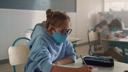 Girl in face mask learning in classroom. Schoolgirl writing in notebook 
