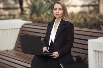 Street style portrait of beautiful business woman in black coat sitting on a bench with laptop