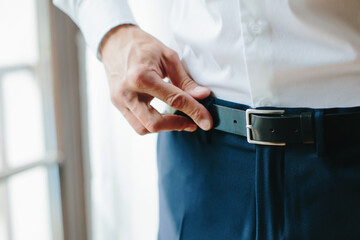 Young man fastens black belt on his trousers, male hands close up. Morning of groom, preparations for wedding. Selective focus.