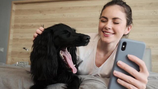 The close-up view of a smiling woman stroking a dog and making a selfie on the phone while sitting on a bed in a room