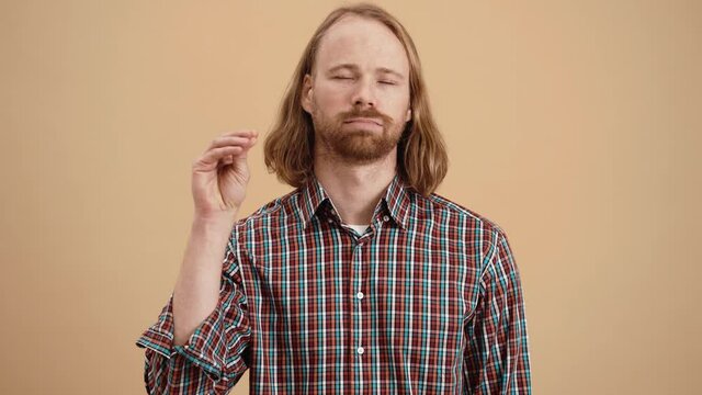 A tired man with long hair in a plaid shirt showing a blah blah gesture with his hand while standing in a beige studio