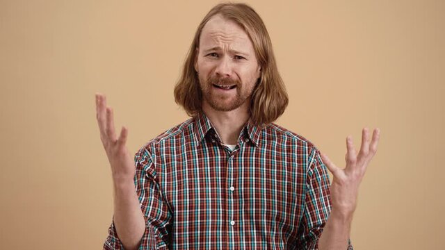 The angry man with long hair in a plaid shirt shouting the what is happening with raised hands while standing in a beige studio