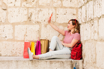 Young woman with colorful shopping bags on the city street makes selfie on the phone. Consumerism, shopping, sales, summer, lifestyle concept.