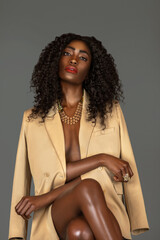 3/4 portrait of a sexy black woman with long curly black hair and beautiful makeup sitting by herself in a studio with gray background just wearing a beige jacket and jewelry.