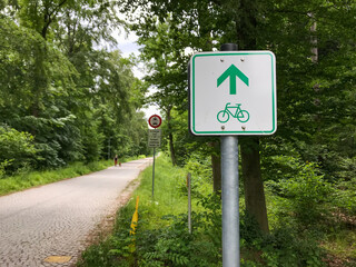 White-green bikeway sign with a straight arrow on a metal pile beside a cobbled road in a forest.
