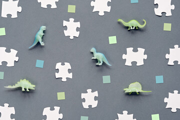 Background with toy dinosaur, white puzzle pieces and green paper squares. Flat lay on grey, silver paper. Concept background for dino birthday decorations.