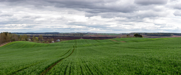 Panorama of a large green hilly agricultural field over a cloudy sky. A village is visible behind the field and peat is being mined.