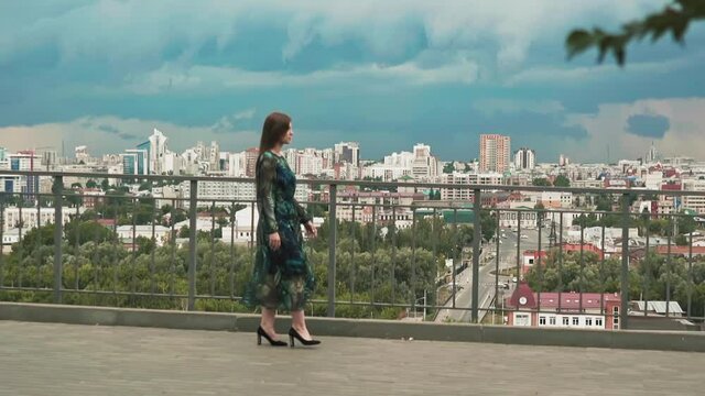 Pretty brunette woman in elegant patterned dress decorated with chiffon walks along empty viewing deck against city and cloudy sky slow motion