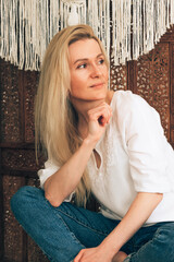 Fototapeta premium A young attractive blonde woman poses in front of a wooden patterned screen decorated with macrame decor.Handmade concept.Home decor.
