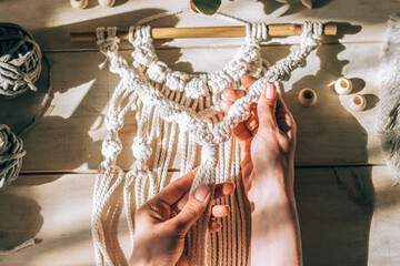 Close up of women's hands weaving macrame from white cotton threads in a home workshop.Home...