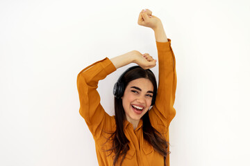 Fototapeta na wymiar Beautiful brunette woman in ocher shirt and black headphones listens to music and smiling on white background. Pretty girl dancing and singing in studio, playing music on her headphones and smiling.