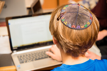 Young Jewish boy wearing colorful Hebrew yarmulke from the back typing on a keyboard at school. - obrazy, fototapety, plakaty