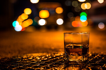 Lights of the city at night through the glass of alcohol, the night avenue with approaching bus. View from the glass level with brandy standing on the wet manhole