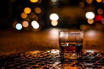 Lights of the city at night through the glass of alcohol, the night avenue with approaching bus. View from the glass level with brandy standing on the wet manhole