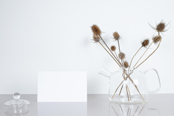Empty greeting card mockup. Glass teapot with dried thistle flowers and beige pillow on light...