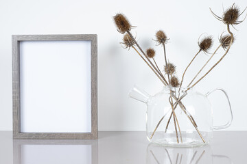Empty wooden frame mockup. Glass teapot and dried thistle flowers on light table.