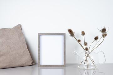Empty wooden frame mockup. Glass teapot with dried thistle flowers and beige pillow on light table.