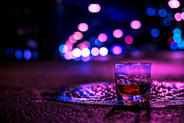Lights of the city at night through the glass of alcohol, the night avenue with headlights of the approaching cars. View from the glass level with brandy standing on the wet manhole