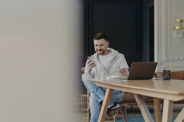 Joyful young male entrepreneur sitting near wooden table and using smartphone while working remotely on laptop from home