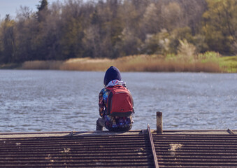Girl in a hood with a red backpack sits by the lake. Beautiful girl from behind on a beautiful landscape background.
