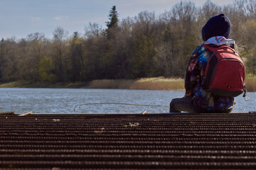 Girl in a hood with a red backpack sits by the lake. Beautiful girl from behind on a beautiful landscape background.