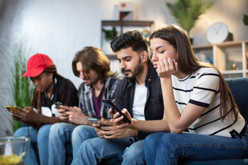 Male and female friends sitting together on sofa and ignoring each other while using personal smartphones. Young diverse people obsessed with modern gadgets.