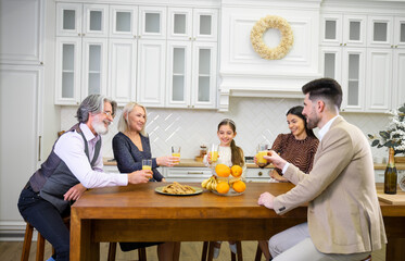 Big happy multi generation family father, mother and grandparents celebrating birthday of cute little girl, clinking glasses with orange juice while sitting at table together in kitchen at home