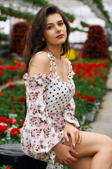 Naklejka premium Attractive young woman in stylish summer dress posing among colorful flowers at orangery. Female model with dark curly hair sitting at looking at camera. 