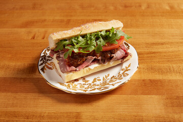 Roast beef sandwich served on an white ornate plate with gold details on a wooden surface