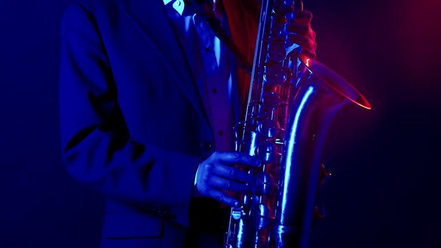 Saxophone is played by male musician a jacket, shirt and hat in a studio in red and blue lighting. Saxophonist performs alone the studio. Concept of musical performance.