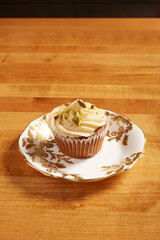 Single carrot cake cupcake served on a white plate with ornate gold details on a wooden surface