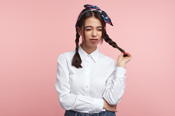 Girl looks unhappily at camera, holds her ponytail against pink background