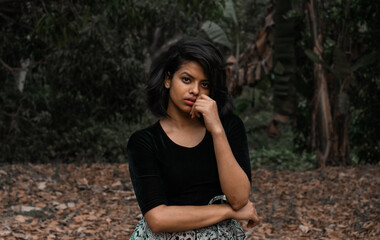 Mid close up of a young woman with short black hair and natural makeup wearing a black top posing in a seductive manner with one hand on her lips in a dark grungy forest