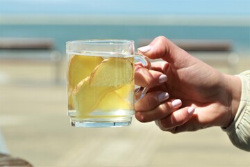 Ginger tea with slices of ginger hold by a hand in front of a beach