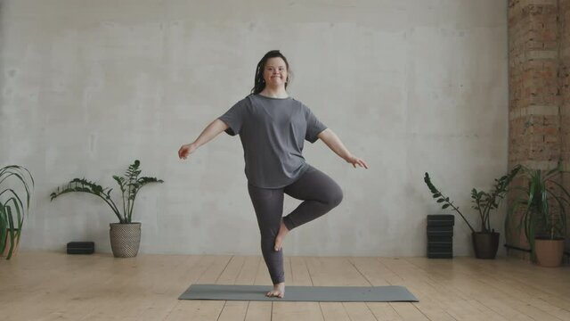 Full shot of young smiling woman with down syndrome standing in yoga pose and looking at camera at bright loft-style yoga studio. Concept of sports motivation for disabled people