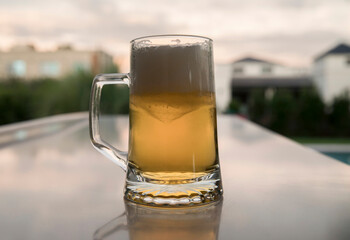 Summer vibes. Closeup view of cold beer mug on the table. The foam, golden liquid and glass on the table in the garden at sunset.
