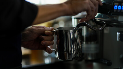 Close-up shot of stainless cup holding by two hands of barista while pouring hot water from steel luxury coffee making machine with blurred cafe background