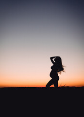 Young adult pregnant woman stands in the field at sunset. Future mom in the twilight. Maternity and fertility concept. Copy space. Pregnancy and motherhhod.