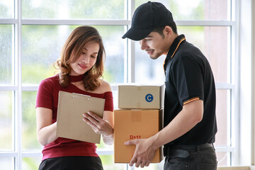 Beautiful Asian female customer signs delivery sheet while receiving packages from smiling handsome Asian courier man. Door to door parcel shipping, e-commerce business concept.