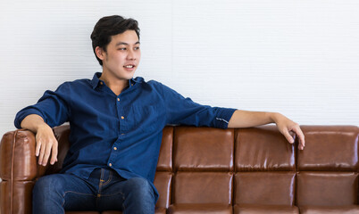 Young man in blue shirt and blue jeans is sitting on a dark brown sofa with smile. He stretch his left arm out for relax.