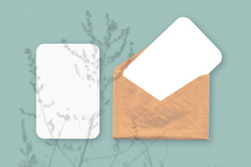 Mockup with an overlay of plant shadows on envelope with two sheets of textured white paper on a green table background. Horizontal orientation