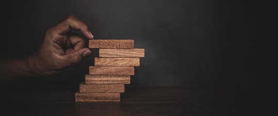Close-up hand is placing wood block tower stacked in stair step with caution to prevent collapse or...