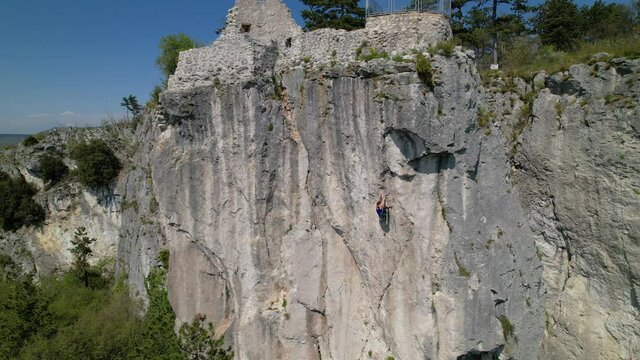 AERIAL: Spectacular rural scenery surrounds athletic young woman learning to top rope climb. Drone point of view of a fit Caucasian female climber top roping up a towering cliff in Crni Kal, Slovenia.