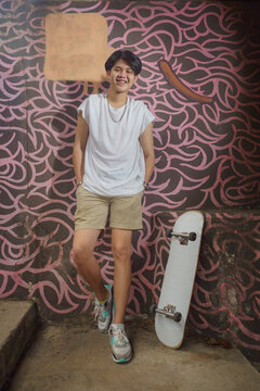 An Asian teenage boy in a white T-shirt and khaki shorts. Smiling standing next to the skateboard