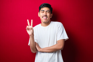 Young handsome asian guy wearing white tshirt standing Doing peace symbol with fingers over face, smiling cheerful showing victory