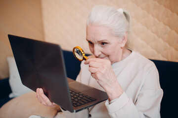 Elderly woman grandmother learns to work at home on laptop computer and internet with magnifier.