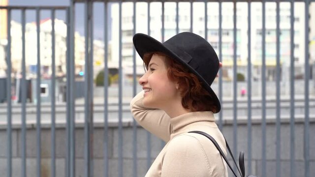 Side view of beautiful smiling red haired woman wearing stylish hat and backpack walking on the street. Travel concept